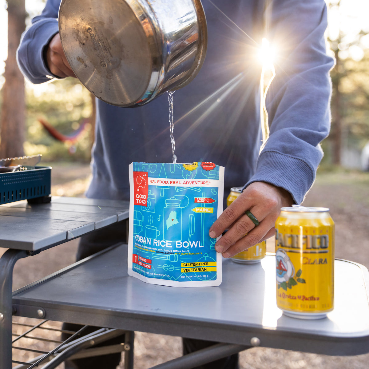 Person pouring water into a package of Good To-Go Cuban Style Rice on a camping table with cans nearby.