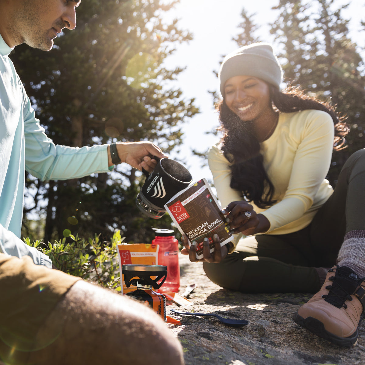 Two people outdoors, one holding good to go pouch while the other pours boiling water to prepare it.