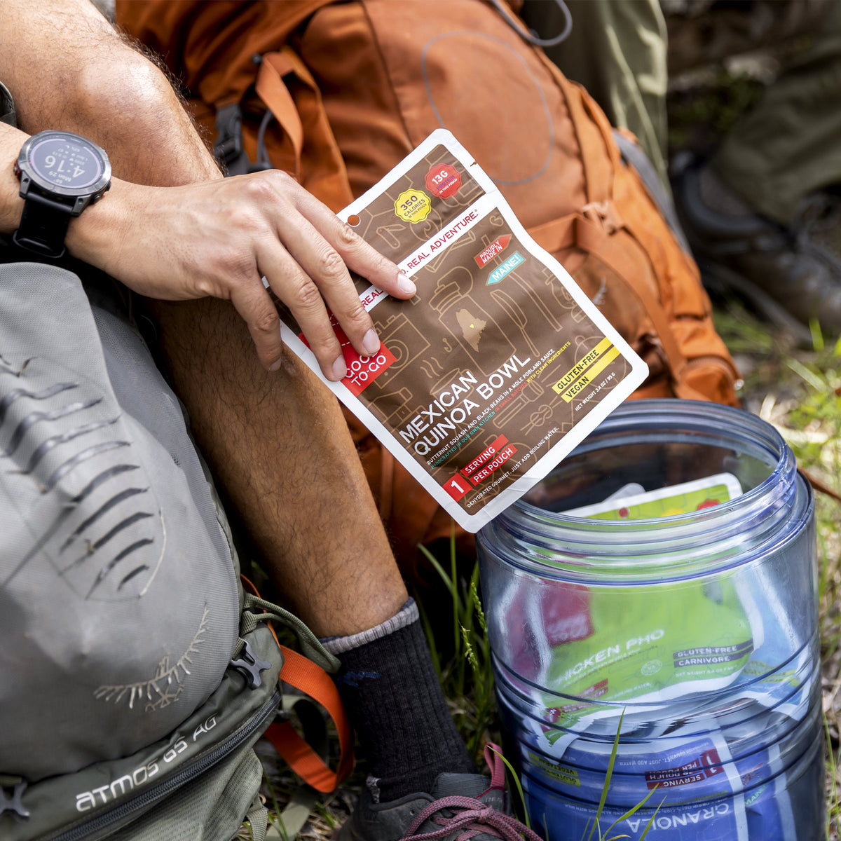 Person holding a package of Mexican Quinoa Bowl over a bear safety storage container with other Good To-Go meals inside it.