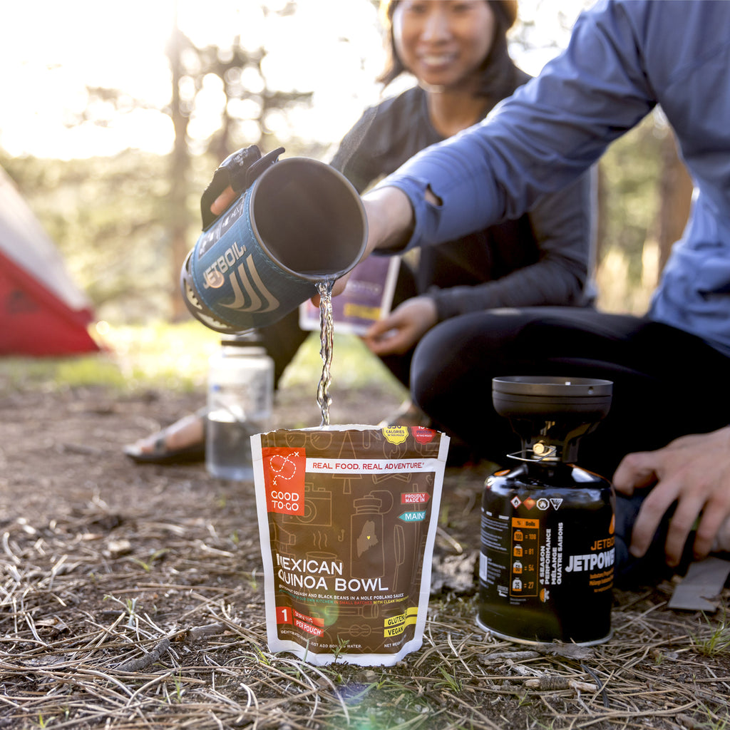 Two people outdoors, one pouring from a jetboil stove into a package of Mexican Quinoa Bowl, with a campfire and tent in the background.