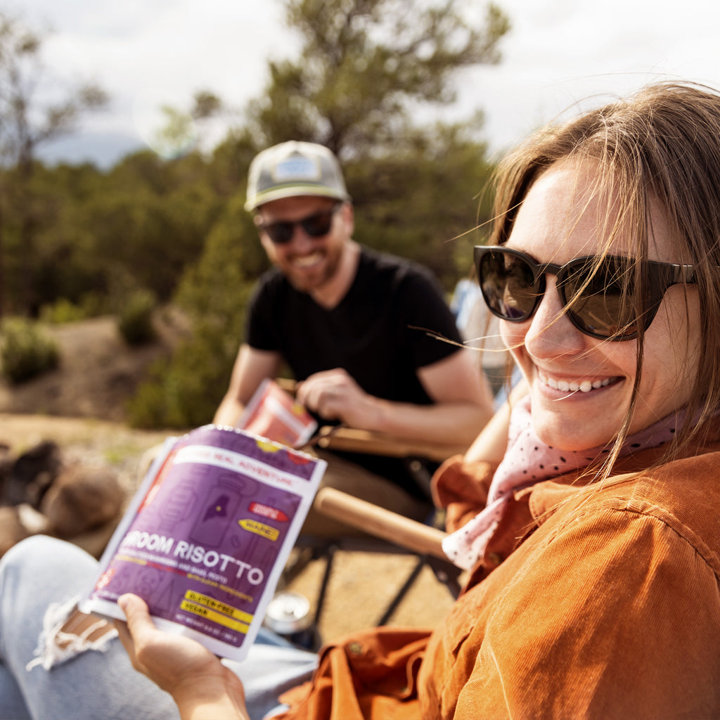 Woman holding a package of Mushroom Risotto while sitting outdoors with a man in the background.