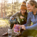 Two people sitting outdoors, one using a Jetboil stove to fill up a mushroom risotto pouch.