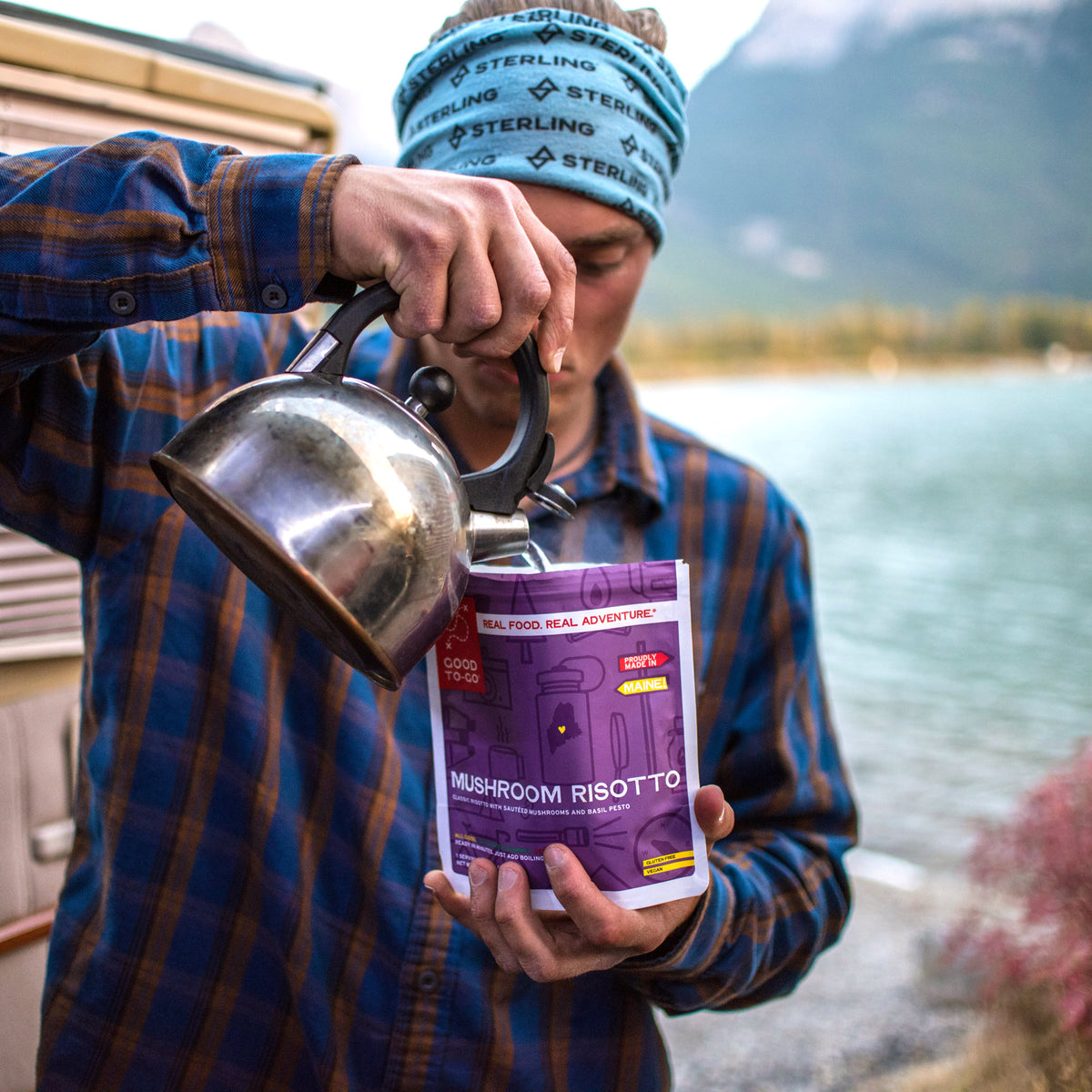 Person outdoors by a lake, holding a pouch of 'Mushroom Risotto' and a filling it up with a kettle.