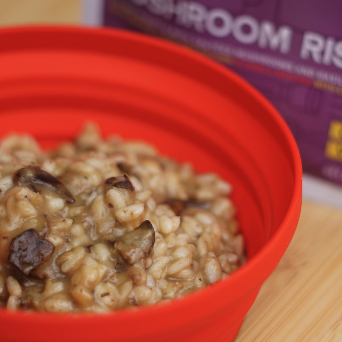 Red bowl filled with mushroom risotto on a wooden surface, with a pouch of mushroom risotto in the background.
