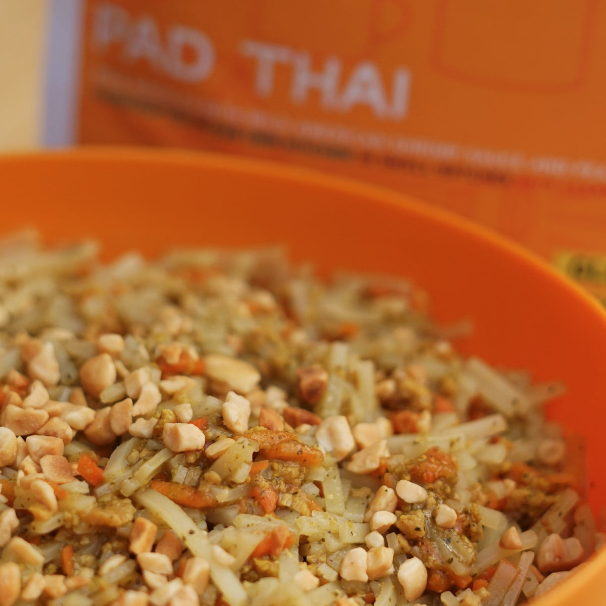 Close-up of Pad Thai in an orange bowl with a blurred 'Pad Thai' box in the background.