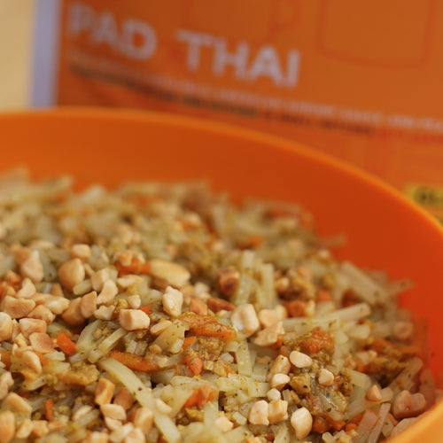 Close-up of Pad Thai in an orange bowl with a blurred 'Pad Thai' box in the background.