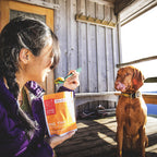 Person holding a package of pad thai with a dog sitting next to them on a wooden deck.