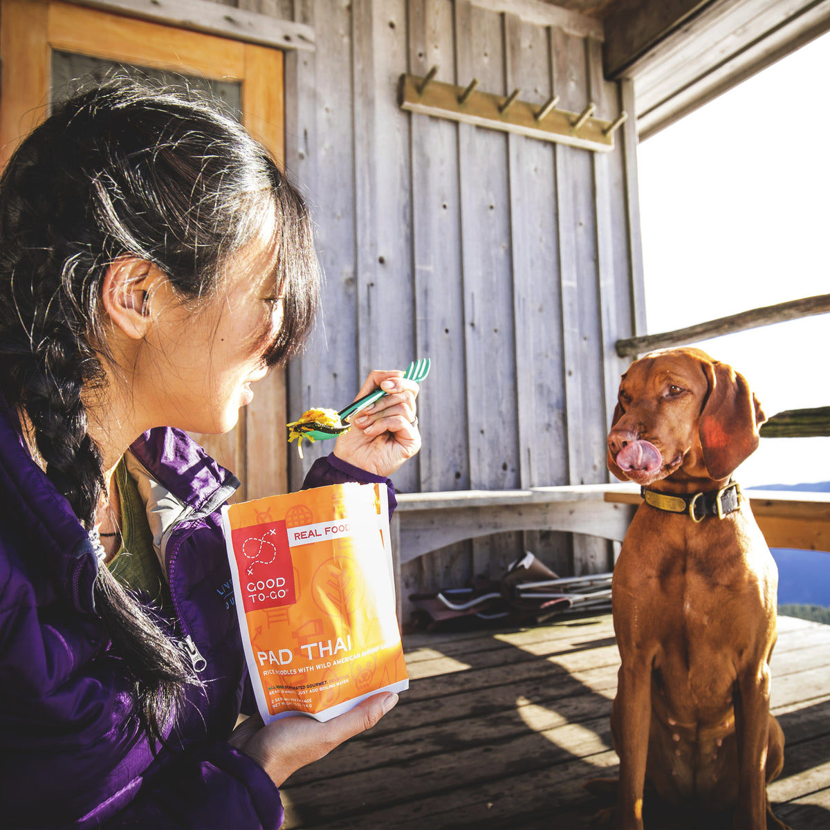 Person holding a package of pad thai with a dog sitting next to them on a wooden deck.