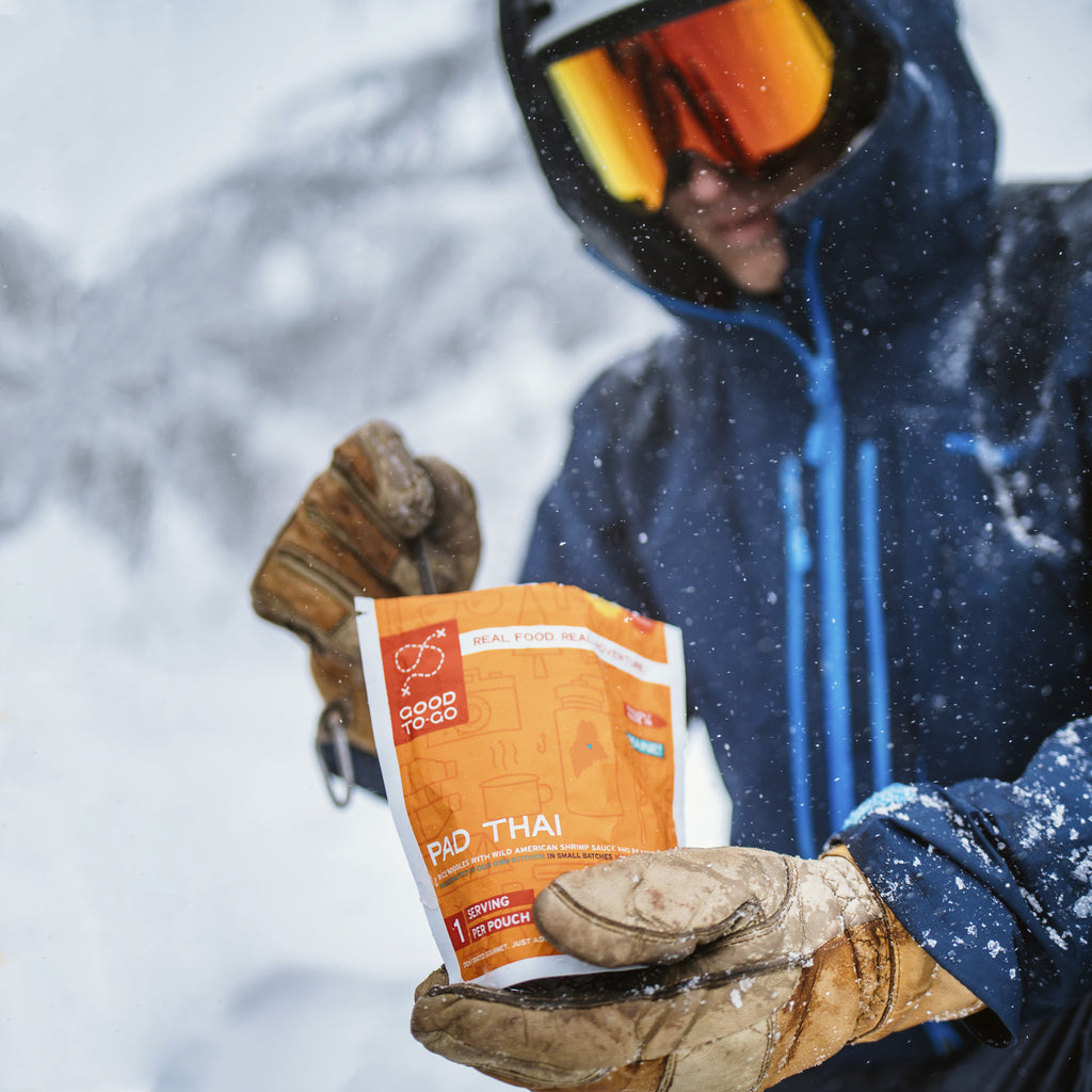 Person in winter gear holding a package of Pad Thai in a snowy landscape