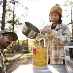 Two people outdoors, one pouring boiling water from a pot into a thai style curry pouch, surrounded by trees.