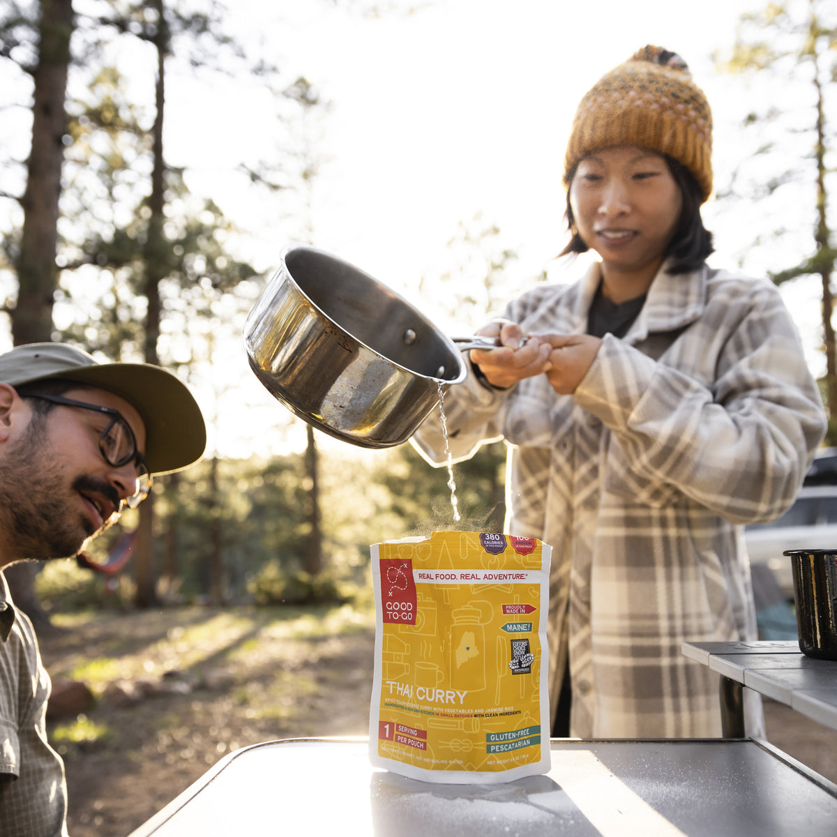 Two people outdoors, one pouring boiling water from a pot into a thai style curry pouch, surrounded by trees.