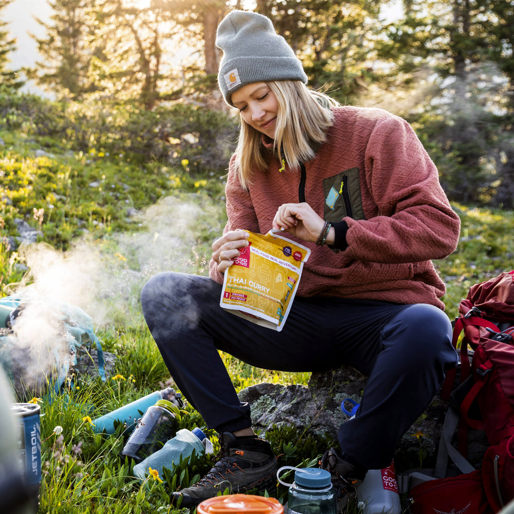 Person opening a Good To-Go Thai Curry package sitting outdoors with a campfire in the background.